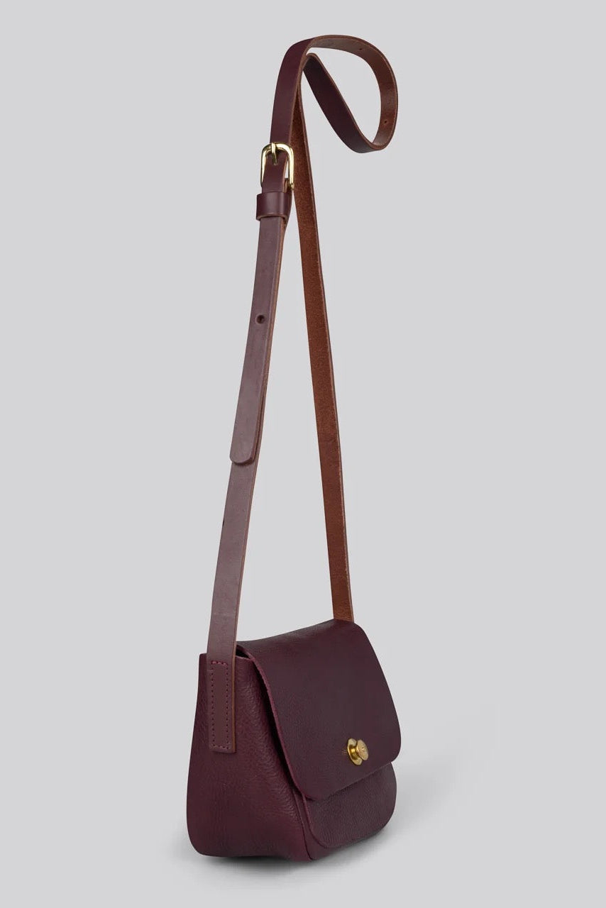 A dark burgundy leather crossbody bag with a matching adjustable shoulder strap and a small gold logo clasp on the front flap, shown against a solid grey background.
