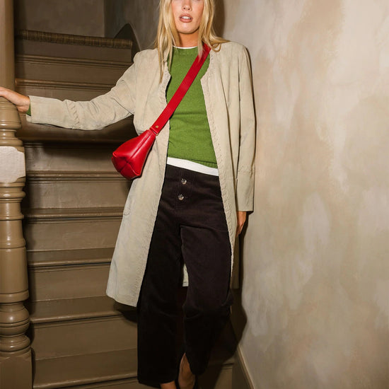 A woman stands on an indoor staircase wearing a light beige coat over a green top, dark wide-leg pants, and black shoes, with a bright red shoulder bag.
