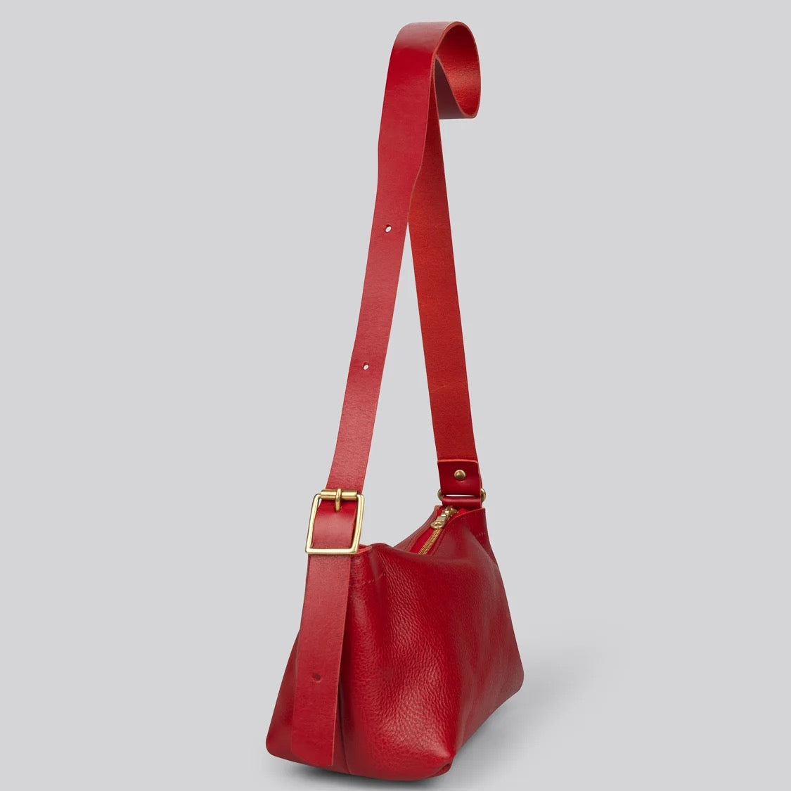 A red pebbled leather shoulder bag with a wide adjustable strap and a gold-tone buckle, shown against a solid gray background.
