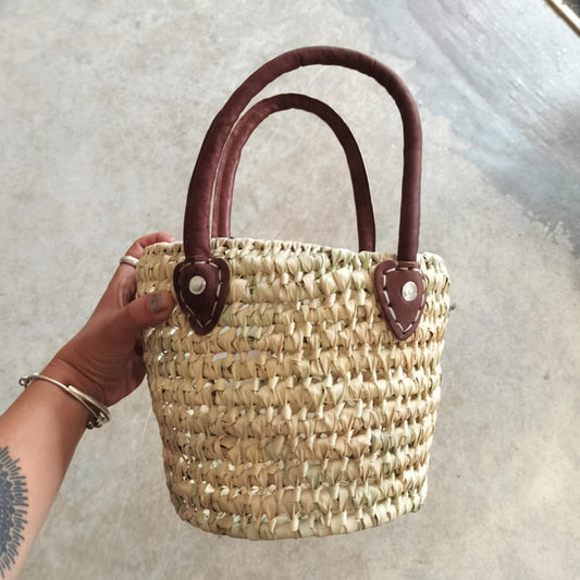 Hand holding a woven basket with brown handles against a neutral background