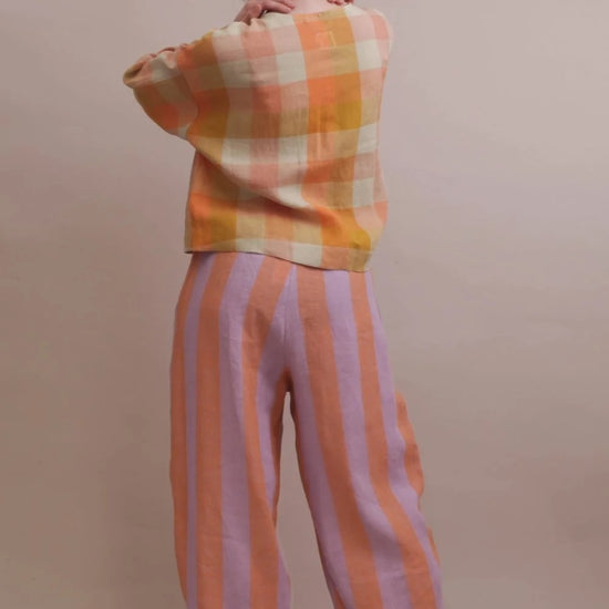 A person shown from the back wearing a cropped plaid top in orange, yellow, and cream, paired with wide-leg striped pants in peach and lavender, against a plain beige studio background.