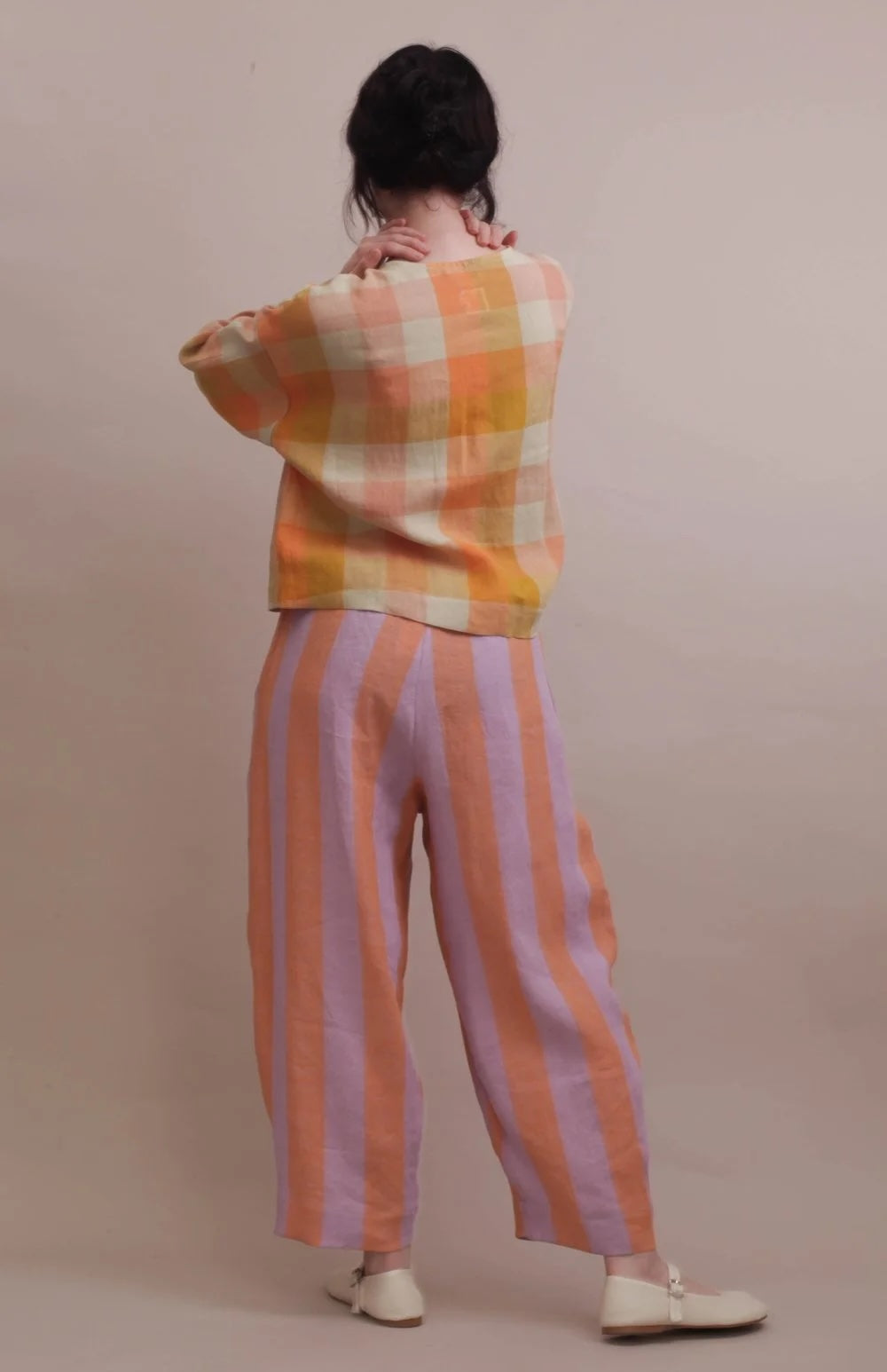 A person shown from the back wearing a cropped plaid top in orange, yellow, and cream, paired with wide-leg striped pants in peach and lavender, against a plain beige studio background.