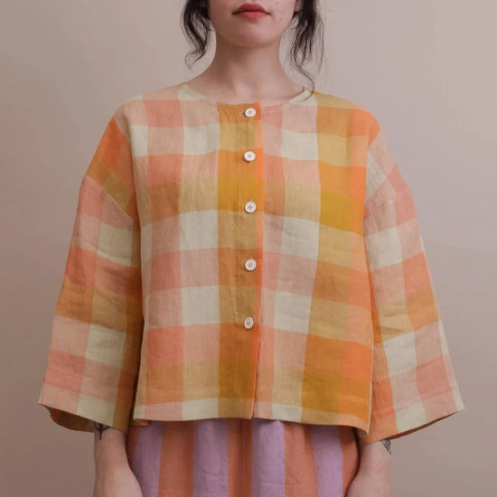 A woman wearing a cropped, three-quarter sleeve plaid top in orange, yellow, and cream, paired with lavender and orange striped pants, standing against a solid beige background.