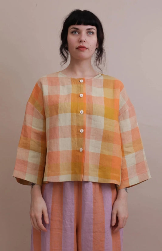 A woman wearing a cropped, three-quarter sleeve plaid top in orange, yellow, and cream, paired with lavender and orange striped pants, standing against a solid beige background.