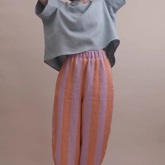 A model wearing a light blue oversized short-sleeve top and wide-leg striped pants in orange and lavender, standing against a solid beige studio background.