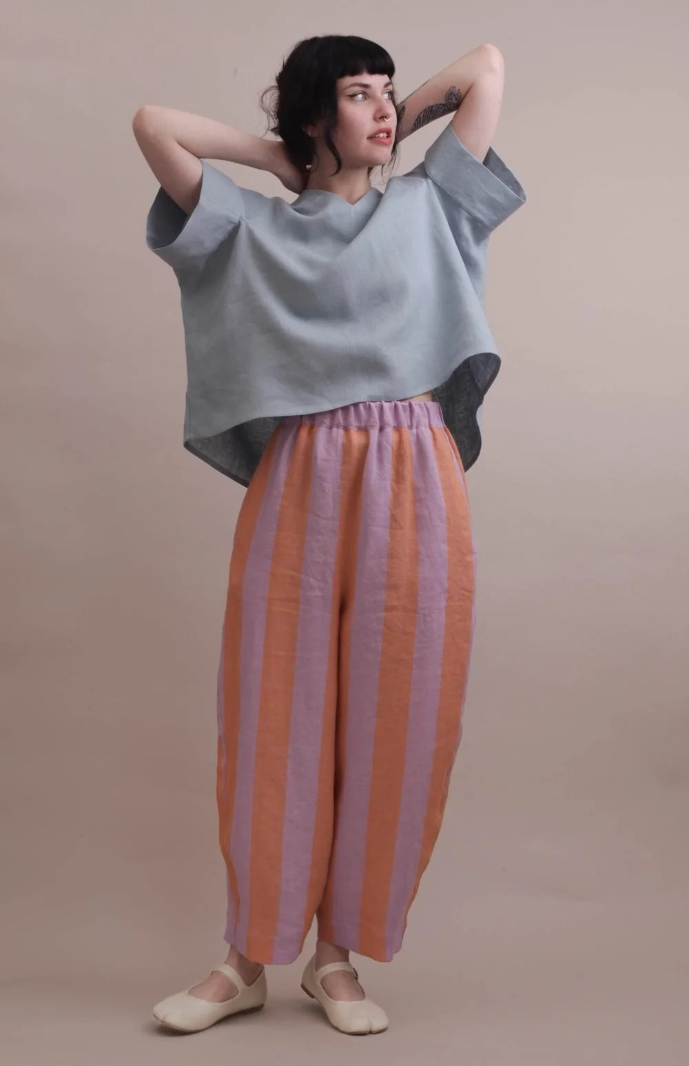 A model wearing a light blue oversized short-sleeve top and wide-leg striped pants in orange and lavender, standing against a solid beige studio background.