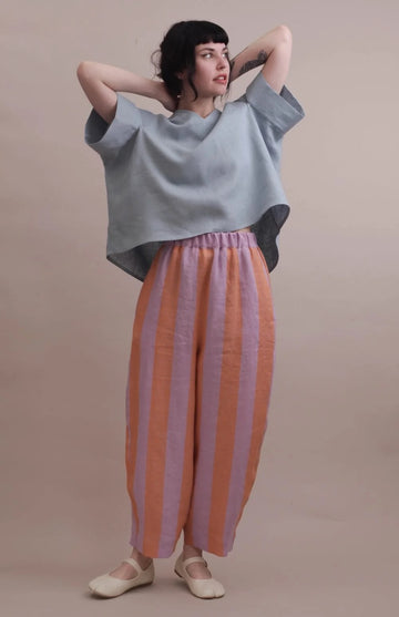A model wearing a light blue oversized short-sleeve top and wide-leg striped pants in orange and lavender, standing against a solid beige studio background.