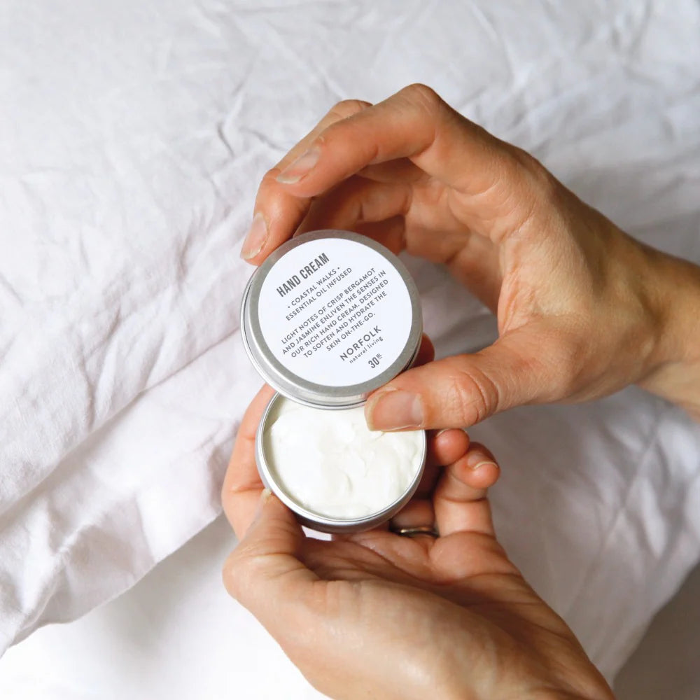 Hand holding a small tin container of hand cream with a white label on a white background