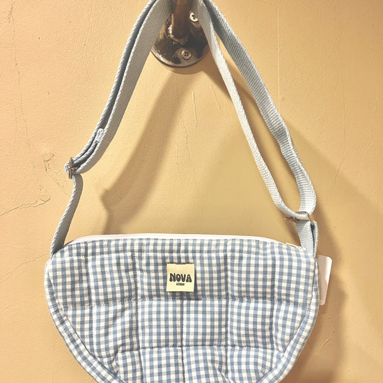 A small quilted crescent-shaped pouch with a light blue and white gingham pattern, featuring a white adjustable strap and a small 'NOVA' brand label. The pouch is hanging from a metal hook against a beige wall.