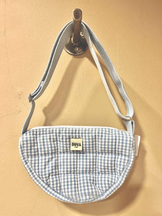 A small quilted crescent-shaped pouch with a light blue and white gingham pattern, featuring a white adjustable strap and a small 'NOVA' brand label. The pouch is hanging from a metal hook against a beige wall.