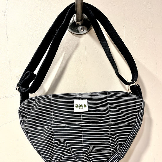A crescent-shaped shoulder bag with a black and white striped pattern, hanging from a silver wall hook. The bag features a white rectangular label with the brand name 'NOVA' printed on it.