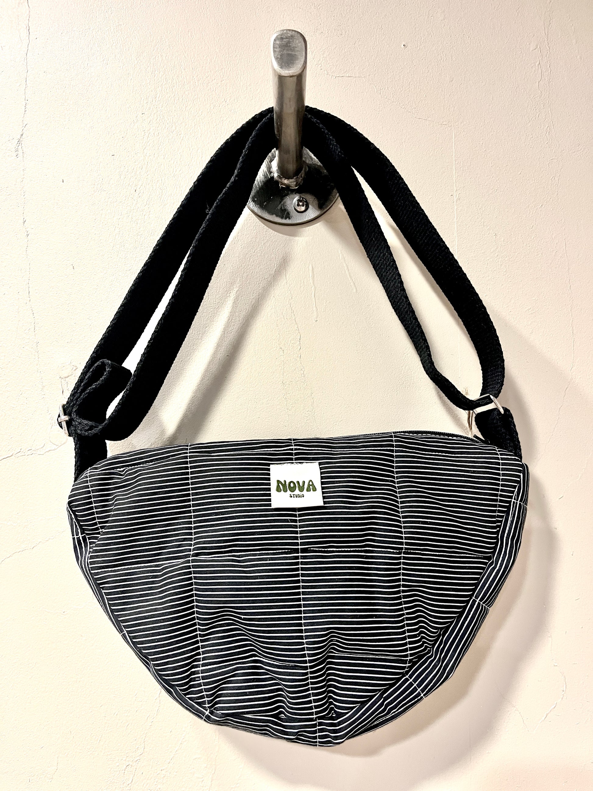 A crescent-shaped shoulder bag with a black and white striped pattern, hanging from a silver wall hook. The bag features a white rectangular label with the brand name 'NOVA' printed on it.