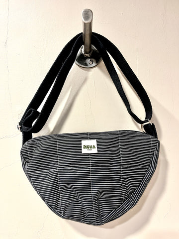 A crescent-shaped shoulder bag with a black and white striped pattern, hanging from a silver wall hook. The bag features a white rectangular label with the brand name 'NOVA' printed on it.