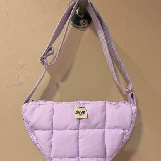 A light purple quilted shoulder bag with a matching fabric strap, hanging from a metal wall hook. The bag features a small cream-colored rectangular patch on the front with the brand name 'NOVA' printed on it, set against a plain beige wall.