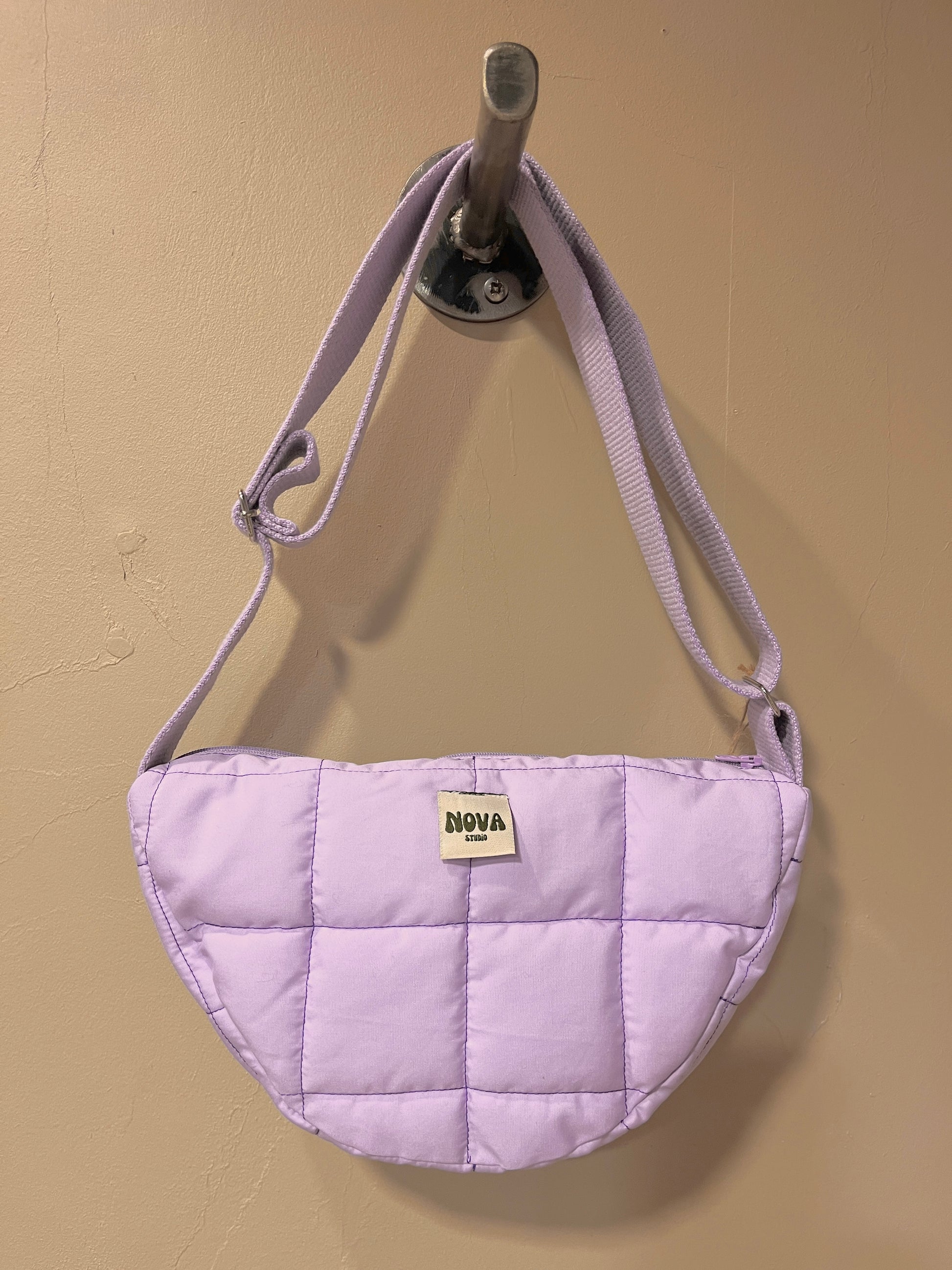A light purple quilted shoulder bag with a matching fabric strap, hanging from a metal wall hook. The bag features a small cream-colored rectangular patch on the front with the brand name 'NOVA' printed on it, set against a plain beige wall.