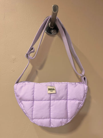 A light purple quilted shoulder bag with a matching fabric strap, hanging from a metal wall hook. The bag features a small cream-colored rectangular patch on the front with the brand name 'NOVA' printed on it, set against a plain beige wall.
