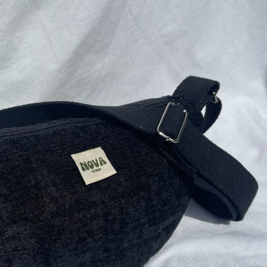 A black textured fabric fanny pack with a wide adjustable strap and a small white 'NOVA London' label, resting on a white fabric background.