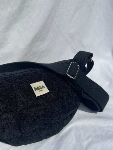 A black textured fabric fanny pack with a wide adjustable strap and a small white 'NOVA London' label, resting on a white fabric background.