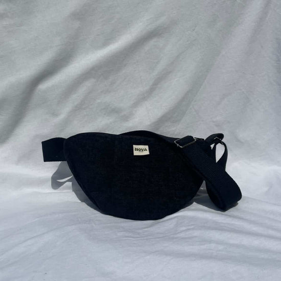 A black fabric cross body bag with a small white brand label reading 'BAGGA' on a wrinkled white fabric background.