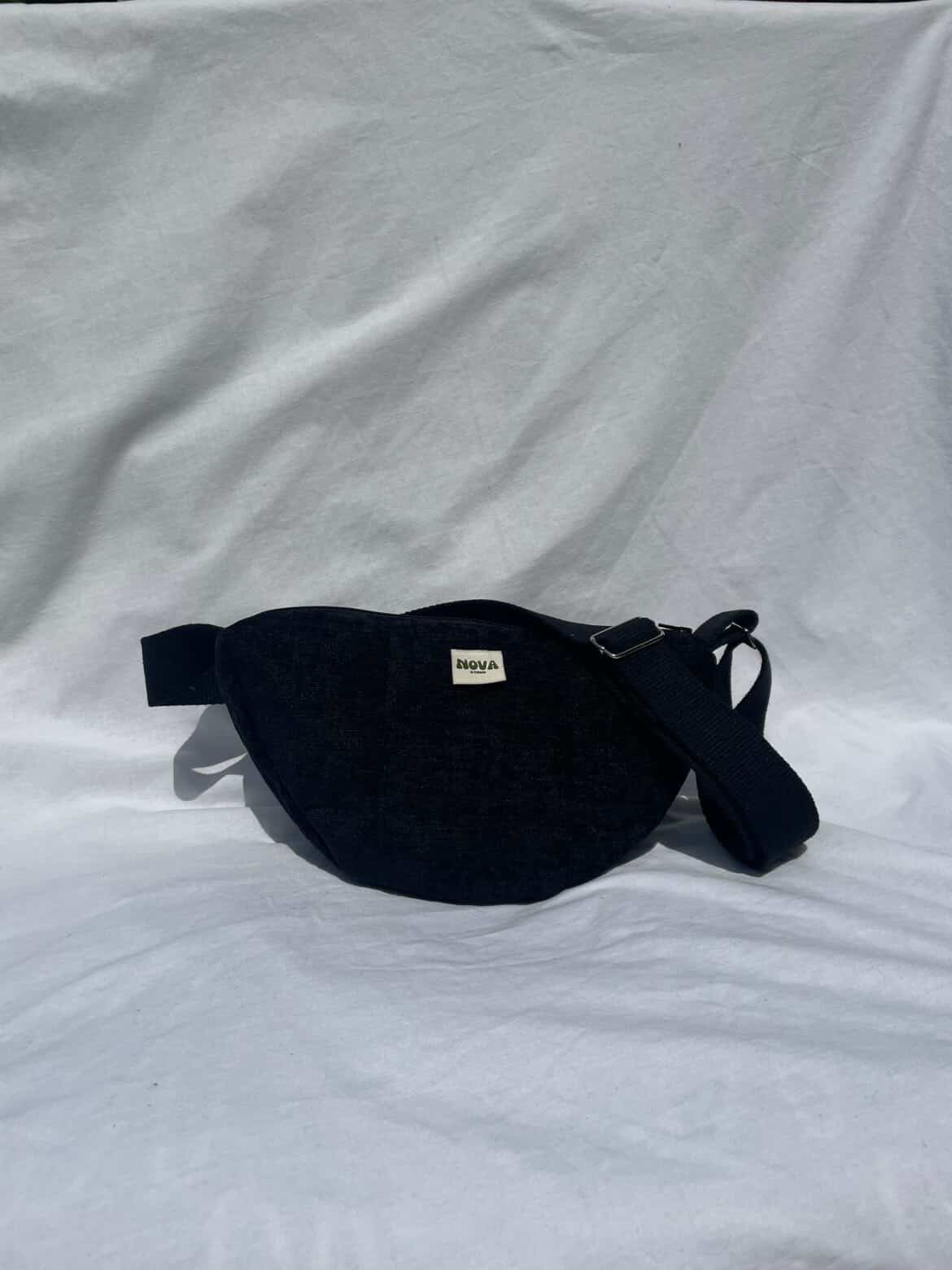 A black fabric cross body bag with a small white brand label reading 'BAGGA' on a wrinkled white fabric background.