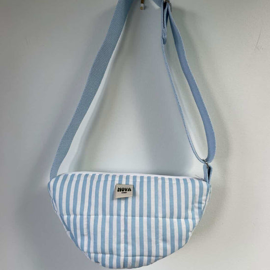 A light blue and white striped crescent-shaped crossbody bag with a matching blue strap, hanging against a plain light grey wall. A small brand label on the front reads 'NOVA'.