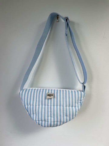 A light blue and white striped crescent-shaped crossbody bag with a matching blue strap, hanging against a plain light grey wall. A small brand label on the front reads 'NOVA'.