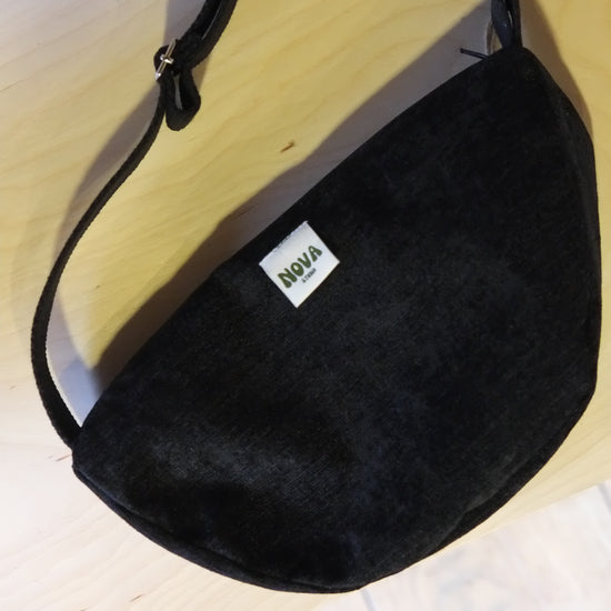 A black suede crescent-shaped shoulder bag with a white rectangular label that reads 'NOVA' in green, resting on a light wood surface.