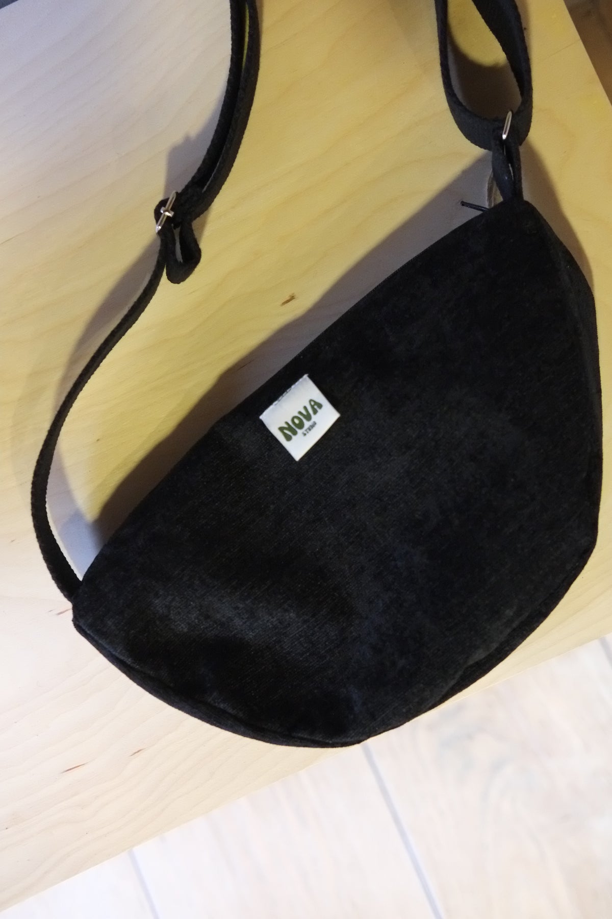 A black suede crescent-shaped shoulder bag with a white rectangular label that reads 'NOVA' in green, resting on a light wood surface.