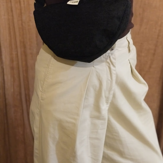 A black textured fabric crossbody bag worn by a person in a dark brown top and cream-colored pants, set against a wooden background.
