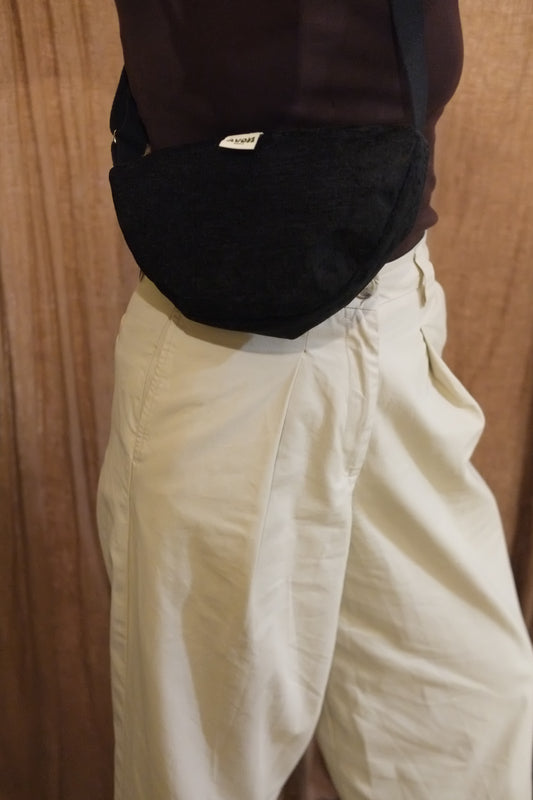 A black textured fabric crossbody bag worn by a person in a dark brown top and cream-colored pants, set against a wooden background.
