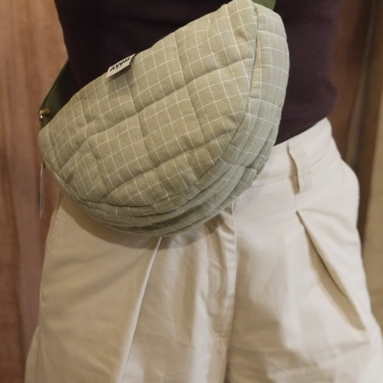 A close-up of a person wearing a quilted, light sage green waist bag with a grid pattern and a green strap, paired with cream-colored pants and a dark brown top.
