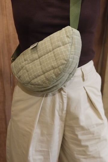 A close-up of a person wearing a quilted, light sage green waist bag with a grid pattern and a green strap, paired with cream-colored pants and a dark brown top.