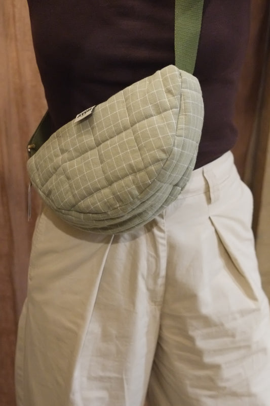 A close-up of a person wearing a quilted, light sage green waist bag with a grid pattern and a green strap, paired with cream-colored pants and a dark brown top.