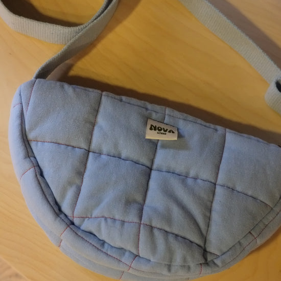 A light blue quilted fabric crossbody bag with a matching strap, resting on a light wood surface. A small fabric label on the front reads 'nova'.