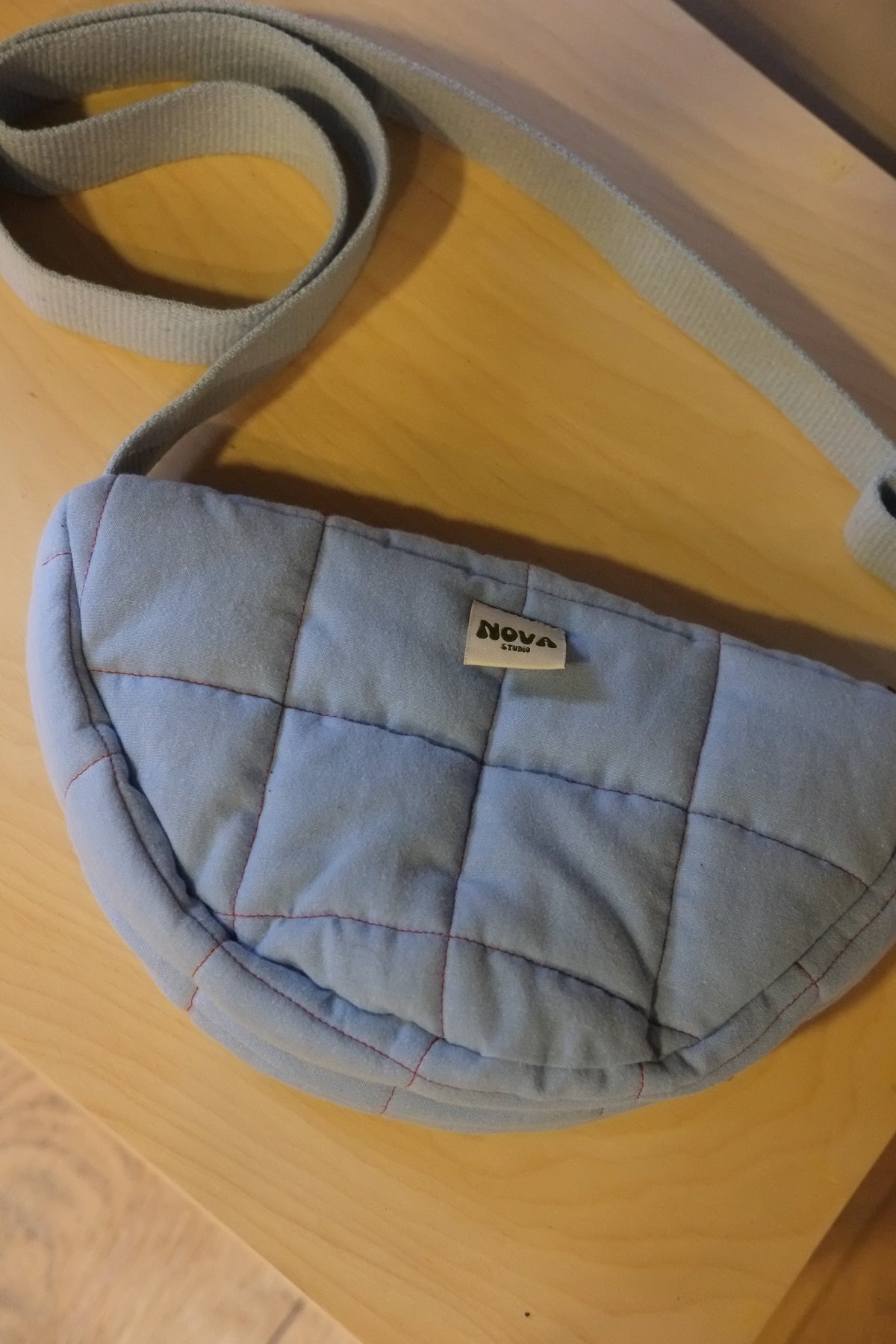 A light blue quilted fabric crossbody bag with a matching strap, resting on a light wood surface. A small fabric label on the front reads 'nova'.