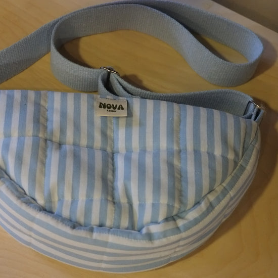 A light blue and white striped quilted pouch bag with a matching fabric strap, featuring a small white 'NOVA' brand tag, resting on a light wood surface.