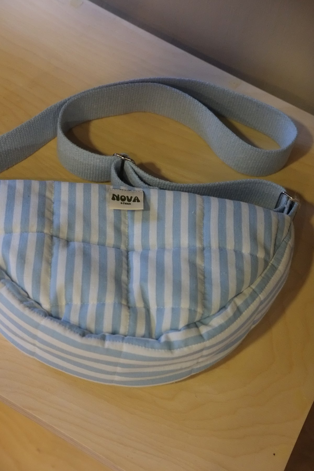 A light blue and white striped quilted pouch bag with a matching fabric strap, featuring a small white 'NOVA' brand tag, resting on a light wood surface.