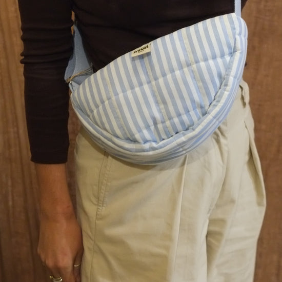 A person wearing a light blue and white striped quilted crossbody pouch bag, styled with a black long-sleeve top and beige trousers, set against a brown textured curtain background.
