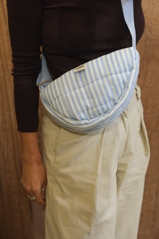 A person wearing a light blue and white striped quilted crossbody pouch bag, styled with a black long-sleeve top and beige trousers, set against a brown textured curtain background.
