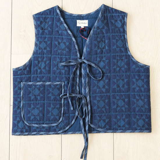 A quilted blue vest with a geometric pattern, featuring a front tie closure and a patch pocket, laid flat on a light wood plank background.