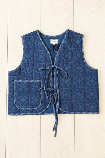 A quilted blue vest with a geometric pattern, featuring a front tie closure and a patch pocket, laid flat on a light wood plank background.