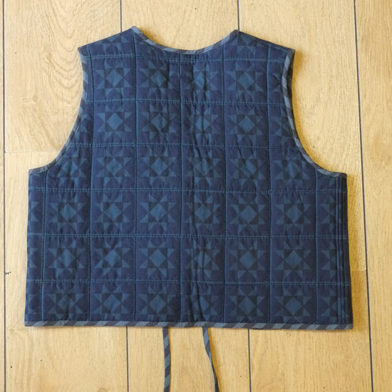 A quilted blue vest with a geometric pattern, featuring a front tie closure and a patch pocket, laid flat on a light wood plank background.