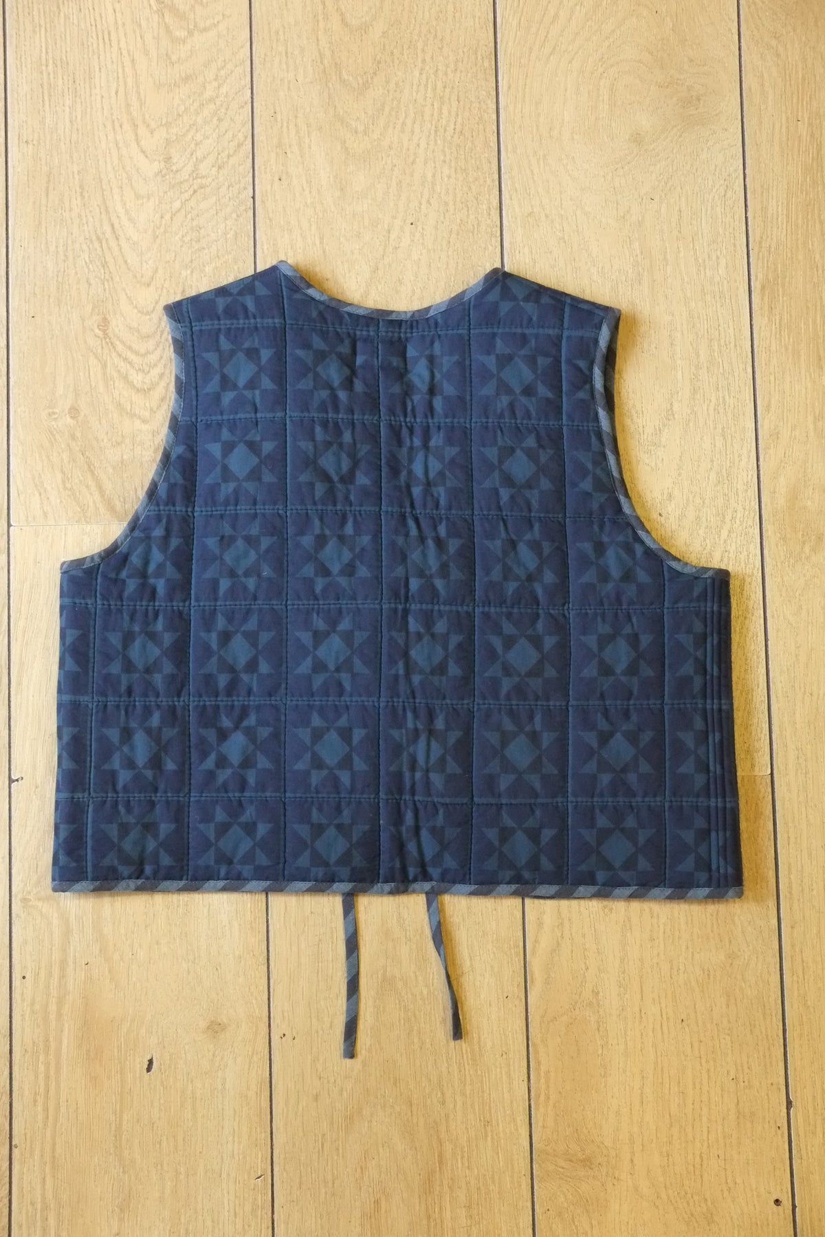 A quilted blue vest with a geometric pattern, featuring a front tie closure and a patch pocket, laid flat on a light wood plank background.