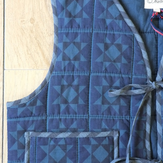 A sleeveless quilted vest in shades of blue with a geometric triangle pattern, laid flat on a light wood floor. The vest features a front pocket and a side tie closure.