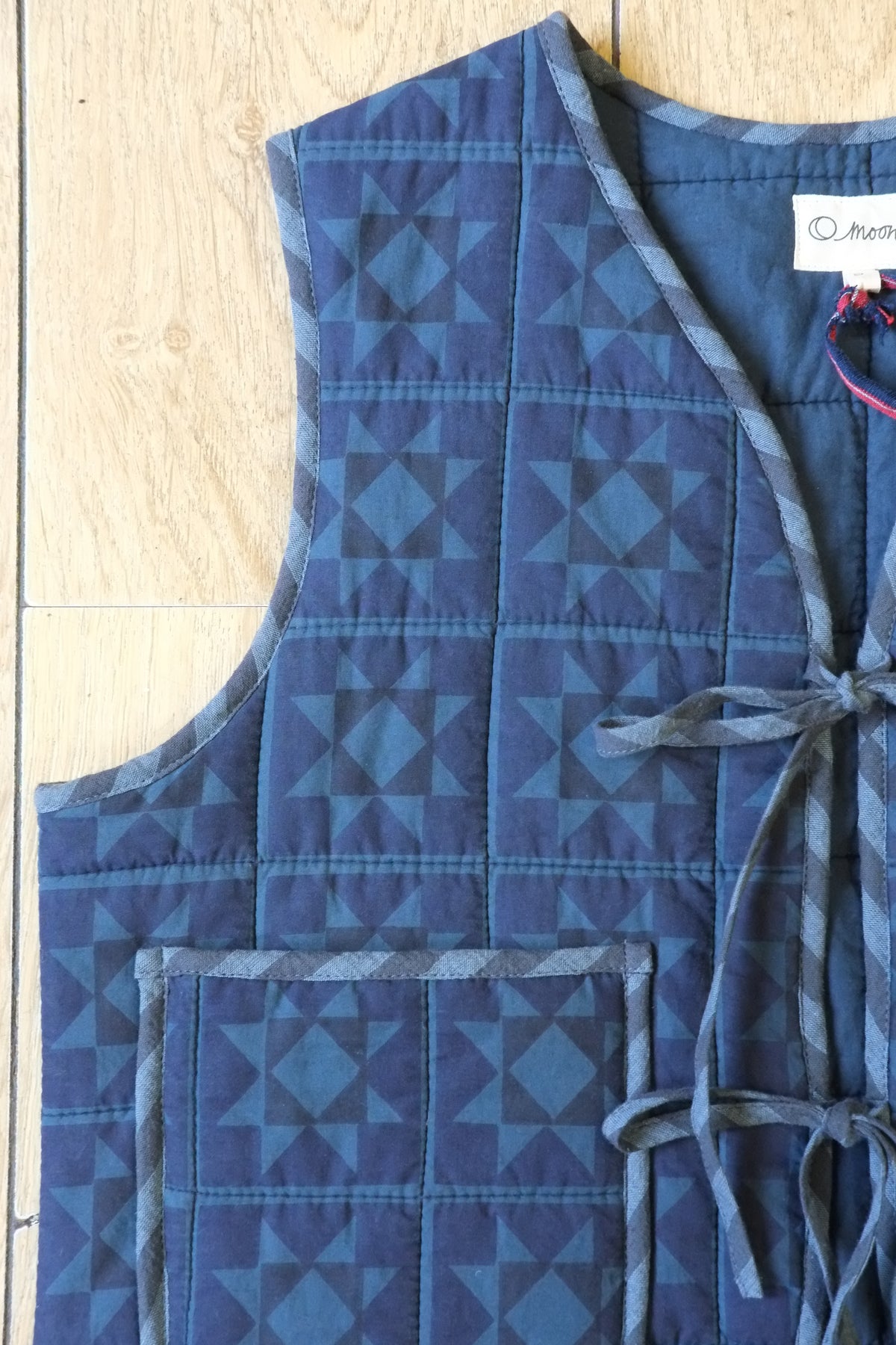 A sleeveless quilted vest in shades of blue with a geometric triangle pattern, laid flat on a light wood floor. The vest features a front pocket and a side tie closure.