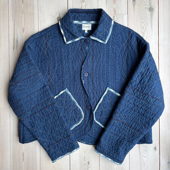 A dark blue quilted jacket with white contrast stitching, laid flat on a light wood plank background.