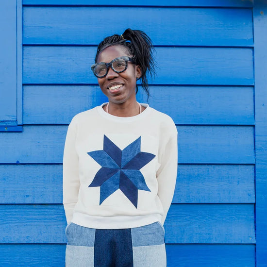 A smiling woman with glasses and her hair in a ponytail stands in front of a bright blue wooden wall, wearing a cream sweatshirt with a large navy starburst quilt pattern on the front.