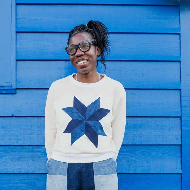 A smiling woman with glasses and her hair in a ponytail stands in front of a bright blue wooden wall, wearing a cream sweatshirt with a large navy starburst quilt pattern on the front.