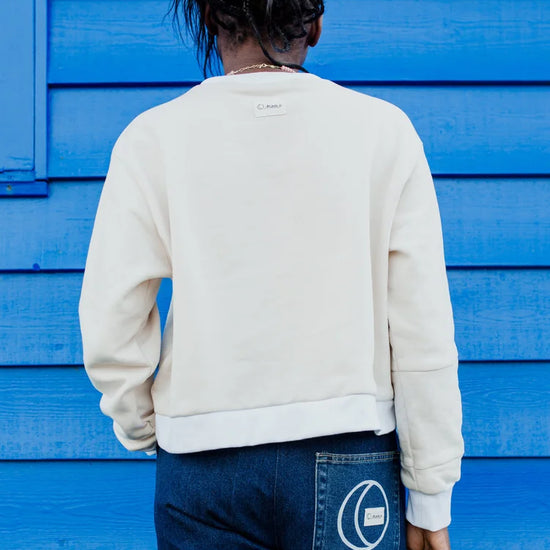 A person viewed from behind wearing a cream-colored crewneck sweatshirt and dark blue jeans, standing in front of a bright blue wooden wall. 