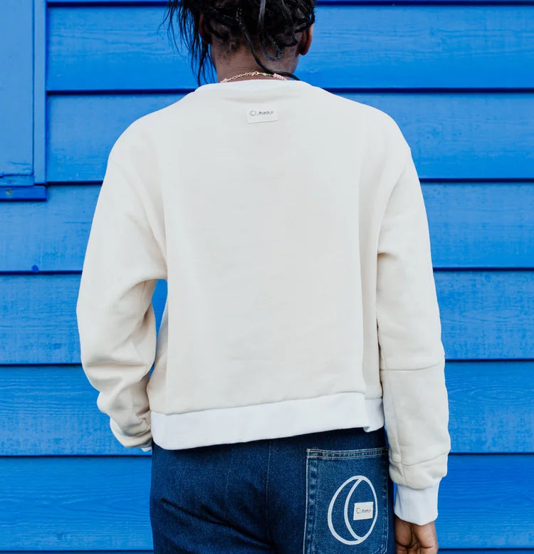 A person viewed from behind wearing a cream-colored crewneck sweatshirt and dark blue jeans, standing in front of a bright blue wooden wall. 